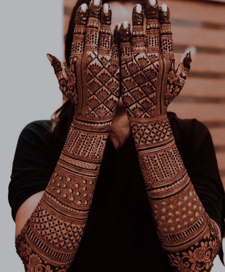 Bride veiling her face behind fully hennaed arms covered in intricate lattice bridal mehandi